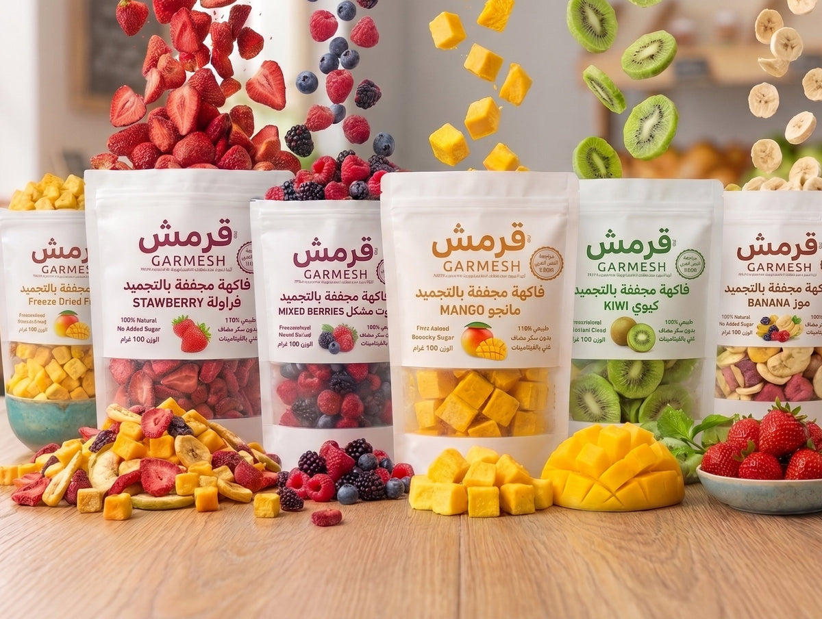 Six stand-up pouches of Garmesh dried fruit mixes in Arabic, arranged on a wooden surface with fresh and dried fruits floating above and scattered around them. The pouches are labeled Strawberry, Mixed Berry, Mango, Kiwi, and Banana, showing colorful dried fruit contents.